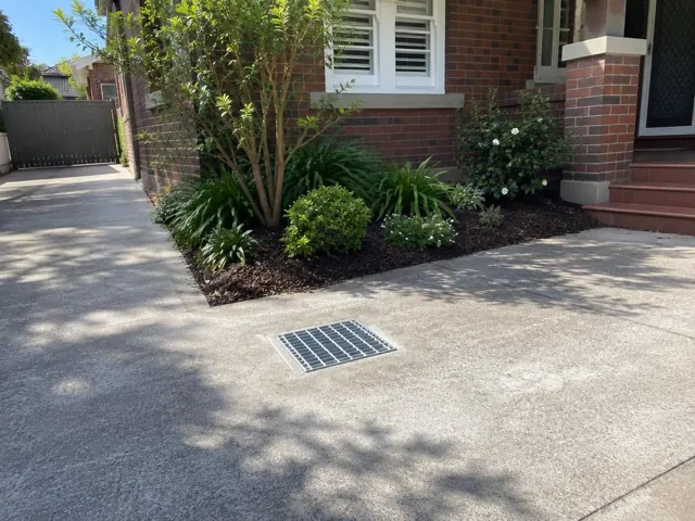 Concrete driveway with a recessed stormwater grate beside a brick house and garden beds in Willoughby